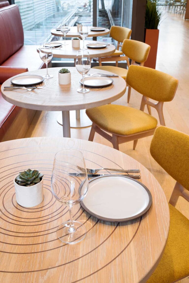 Series of tables set for two at Clay Restaurant, the wood top of the table is engraved with concentric circles, and the chairs have yellow coverings.
