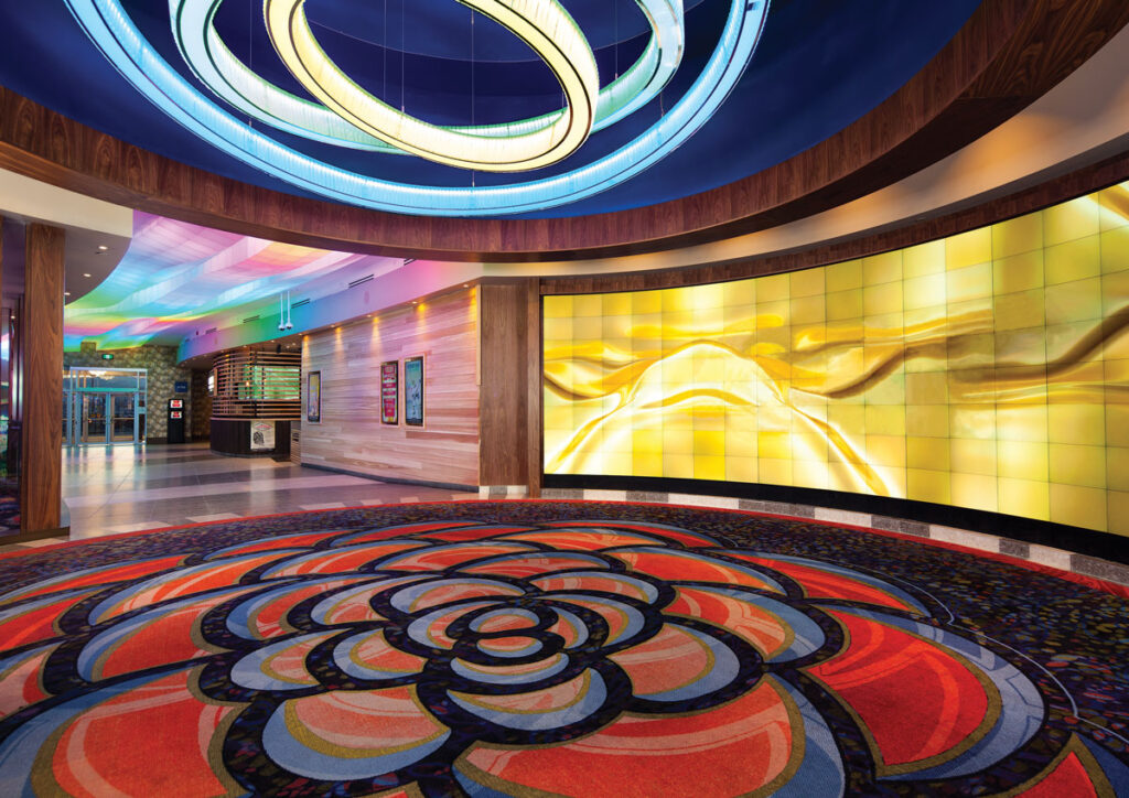 Carpet with a large scale floral pattern in casino lobby with a light fixture of concentric rings is suspended above.