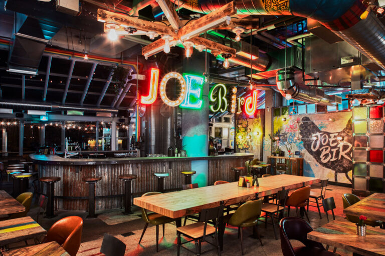 Interior view of Joe Bird restaurant with wood topped tables mismatched chairs, neon sign of restaurant name and open ductwork.