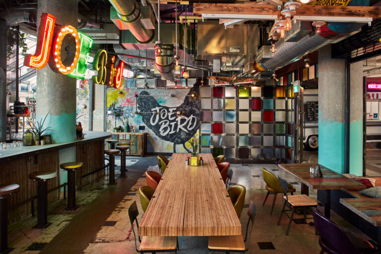 Communal table for 10 at Joe Bird restaurant with wood topped table and mismatched chairs. Decorated ductwork overhead.