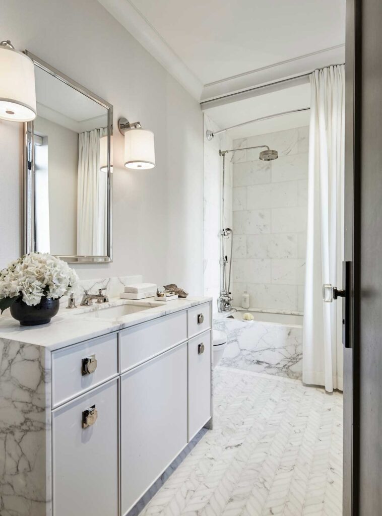 Bright white bathroom with herringbone marble flooring, and white marble topped sink, with a soaker tub and shower on the far wall.