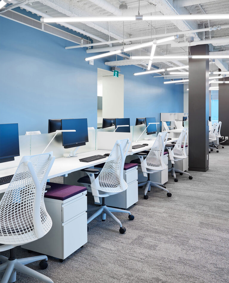 Employee workstations in white with matching seating against a calm blue wall. Modern fluorescent lighting crisscrosses overhead.