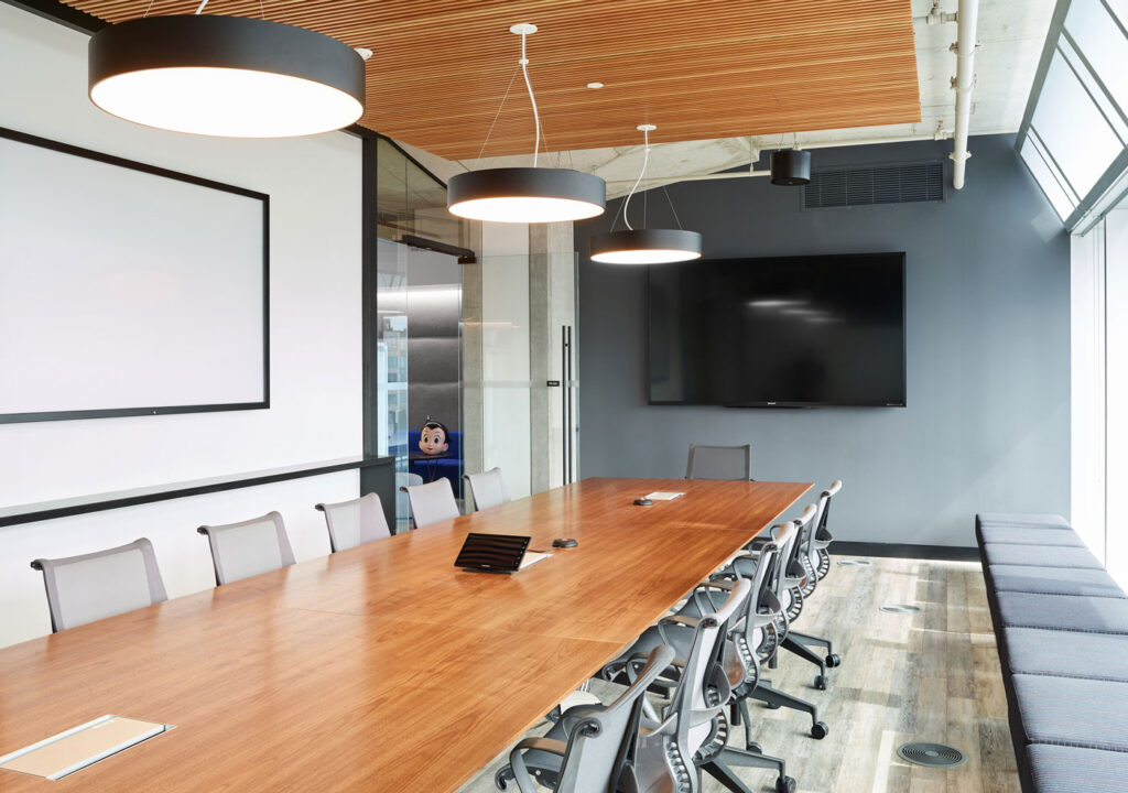 A large meeting room with wood topped conference table surrounded by office chairs and bench seating nearby. Round black pendant lights overhead extend the brand while a tambour wood screen hides acoustical panelling.