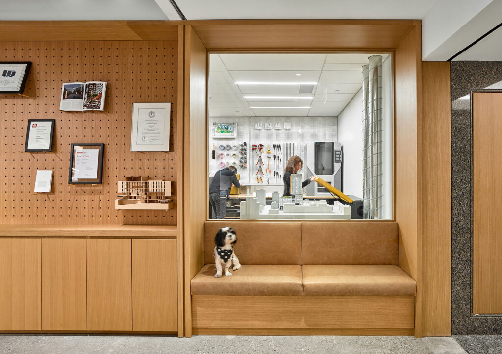 A small black and white dog sits on a maple leather bench seat inset into the wall. The wall behind the seat has a window into an architectural model shop.