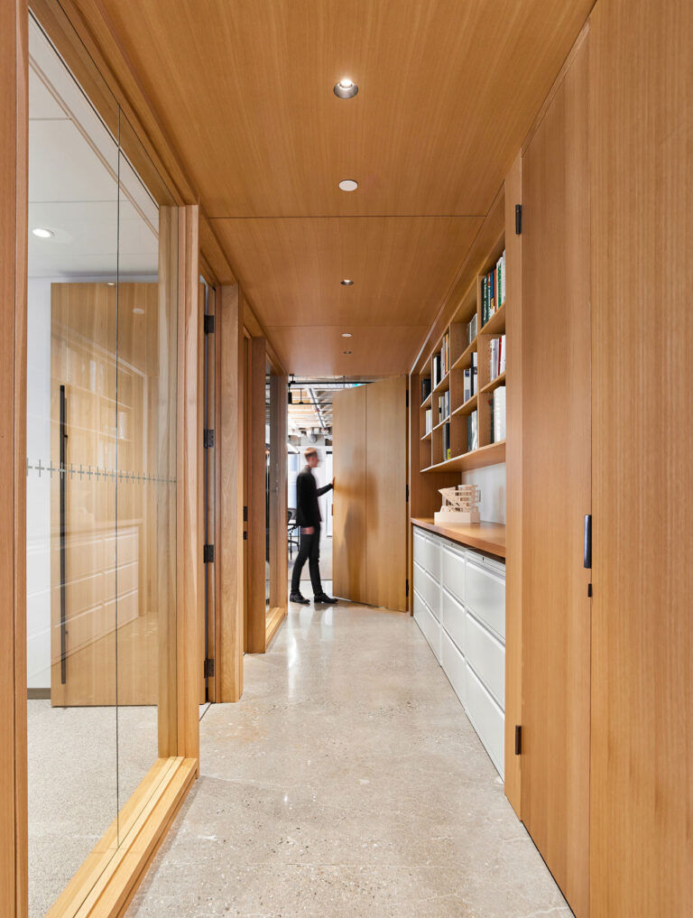 A corridor lined in maple wood with several glass walled meeting rooms opposite shelving and file space.