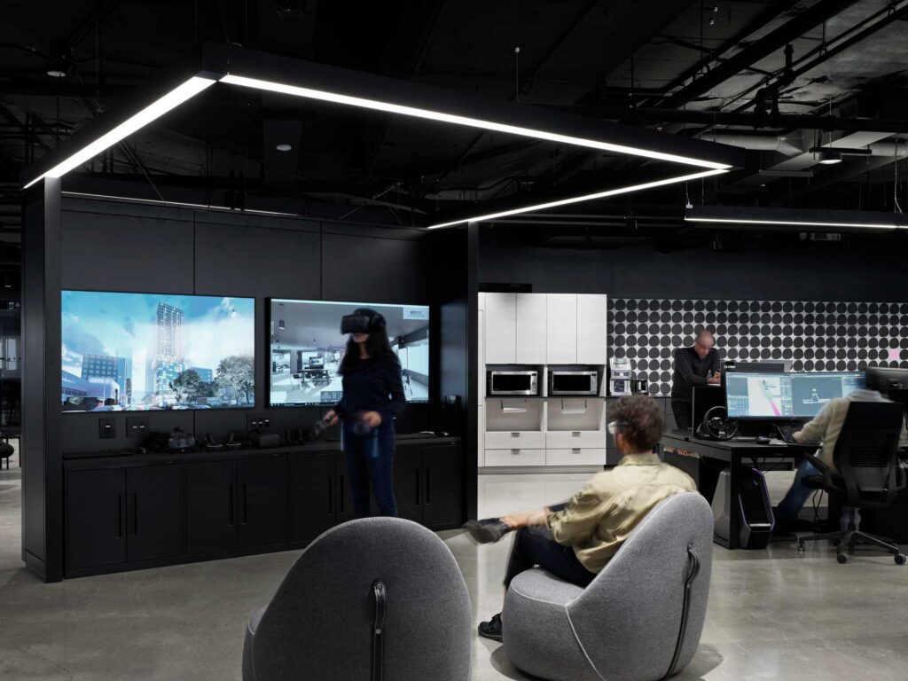 A woman uses a VR headset in the Smart City Sanbox in a room with a black console with TV screens and a polished concrete floor.