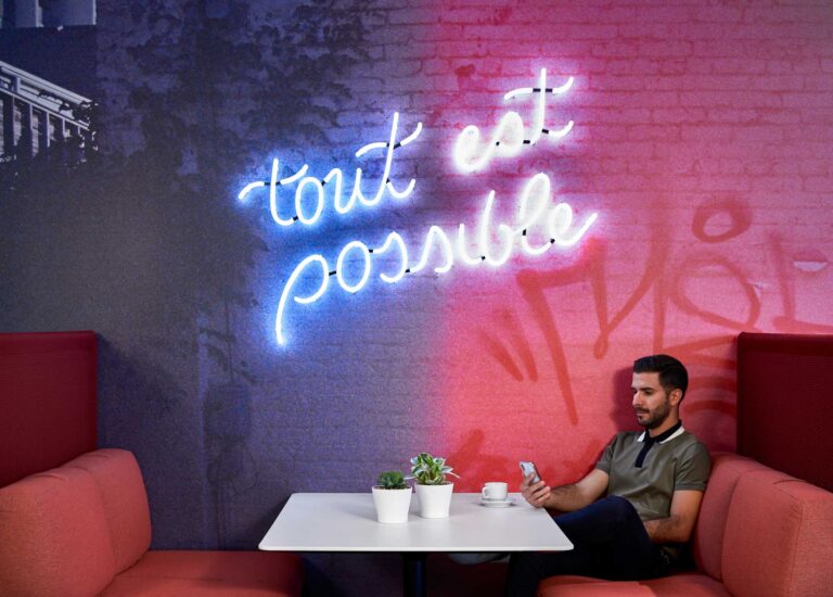 A man looks at his phone while seated at a pink and red booth under a neon sign which reads "Tout est possible".