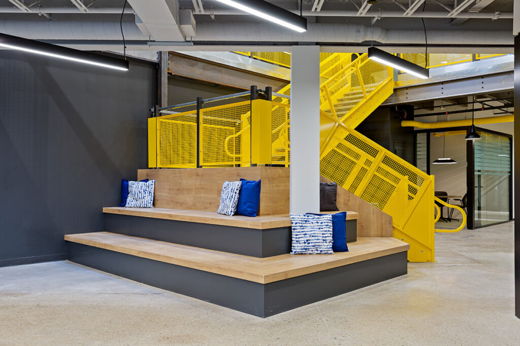 Multi level seating area with pillows and behind it a bright yellow stair case leading to the second floor of the office space