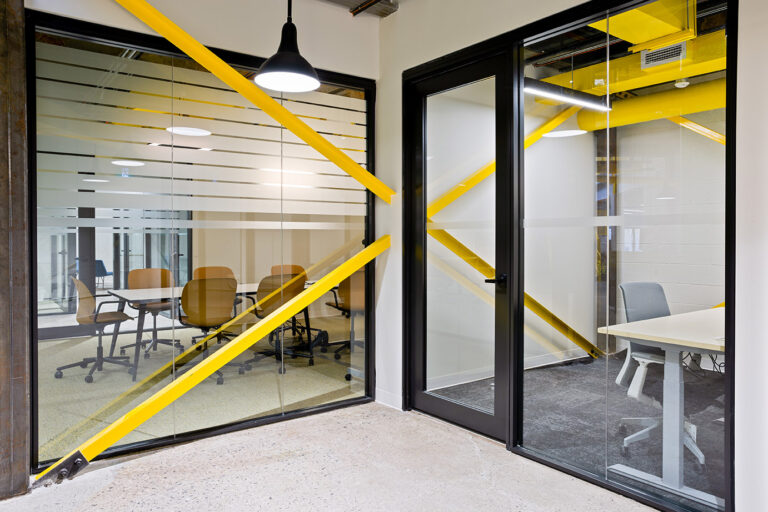 A view of one of the meeting rooms behind a glass wall and next to it an office behind closed glass doors. Connecting them is steel beam X shaped structure in bright yellow color.