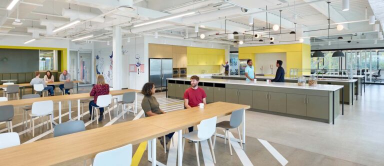The open cafeteria area at Text now has cabinets covered in a soft yellow, olive and wood topped tables surrounded by windows for natural light.