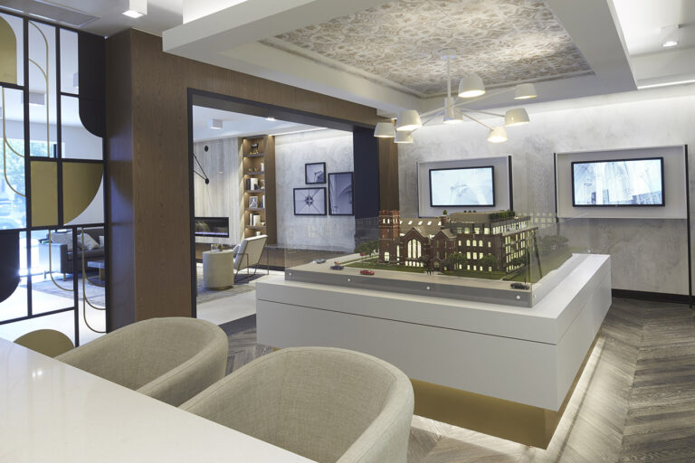 Central area of the presentation center with the large 3D model of the condo building and a large modern white light fixture above. Two monitors on the wall in the back.