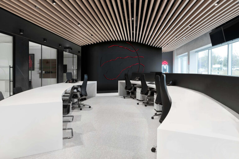 Open concept office space with long white curved desks on either side of the room. Custom ceiling framework in a form of slats leading the eye to the black feature wall branded with a discreet 3D raptors logo outline on the far end of the space.