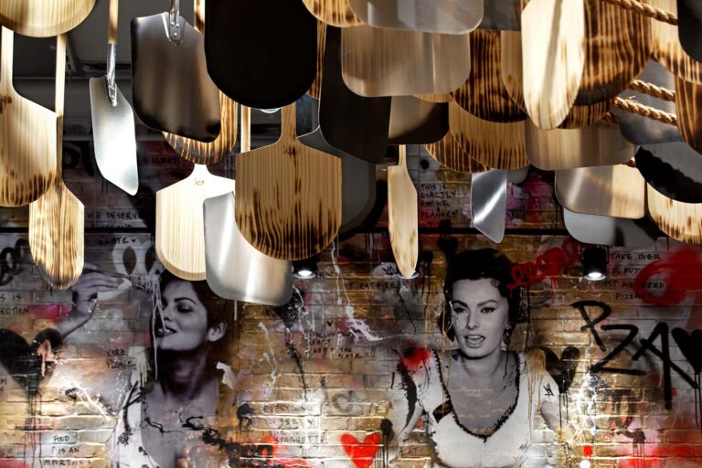 An installation of charred pizza peels is hung from the ceiling of the restaurant against a graffiti inspired mural featured iconic Italian actress Sophia Loren.