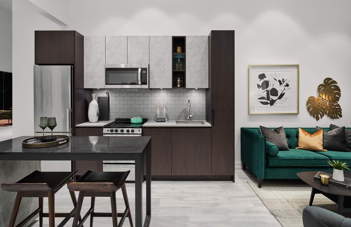 Model kitchen in brown wood and grey color finishes. To the right of the kitchen is a living room and a luxurious emerald green sofa with gold accents.