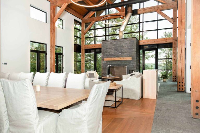 Main living open concept area encased in large windows showing views of the woods on all sides. The seating is concentrated around the huge grey brick fireplace