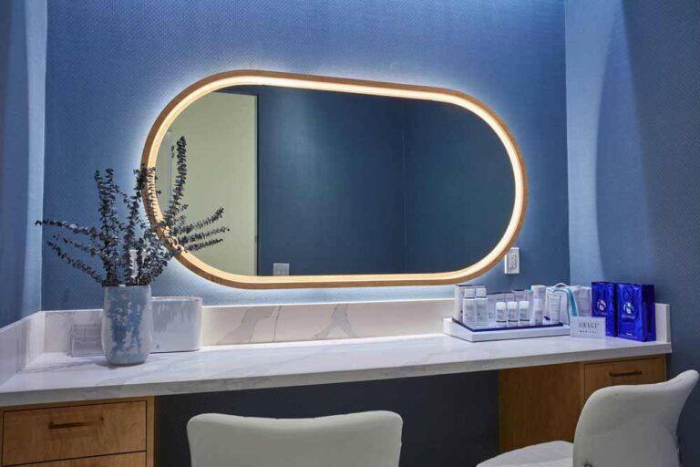 Blue tranquil wall in the make up room, with a beautiful horizontal mirror with rounded edges above a modern marble and wood mix vanity. Some facial products and a flower vase are placed on the vanity top.