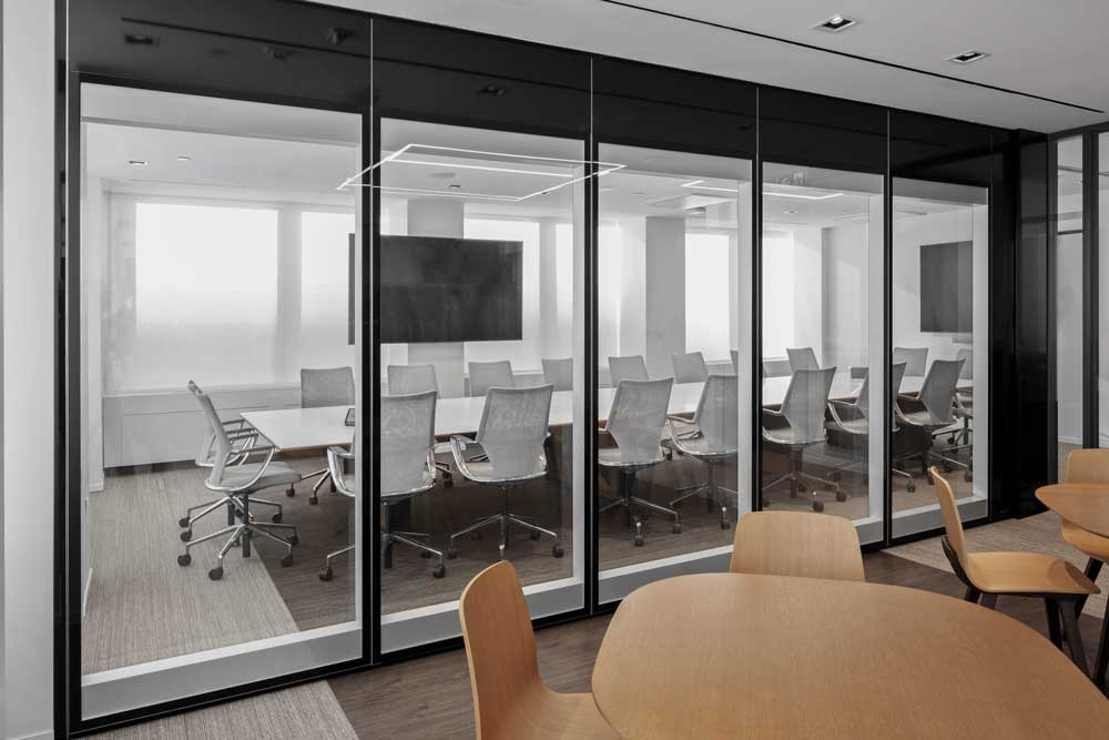 Large long meeting room in neutral color scheme and modern furniture is visible behind a glass wall, looking from a common seating area in wood finish