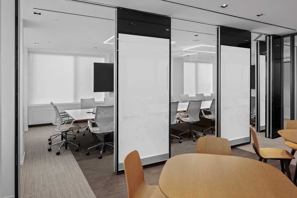 Large long meeting room in neutral color scheme and modern furniture with opened glass wall panels, looking from a common seating area in wood finish