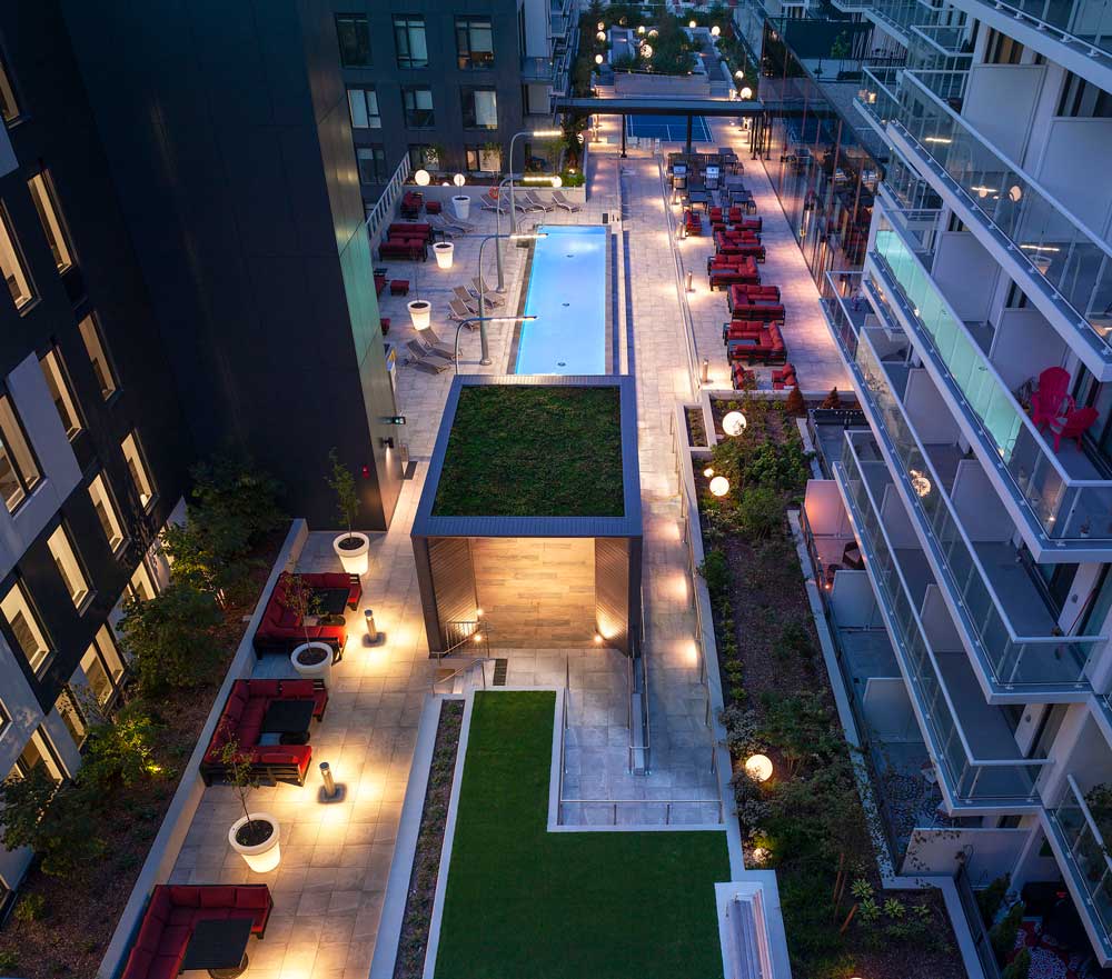View of the long and narrow common outdoor area at night with lit up pool and pop of colour in red patio seating
