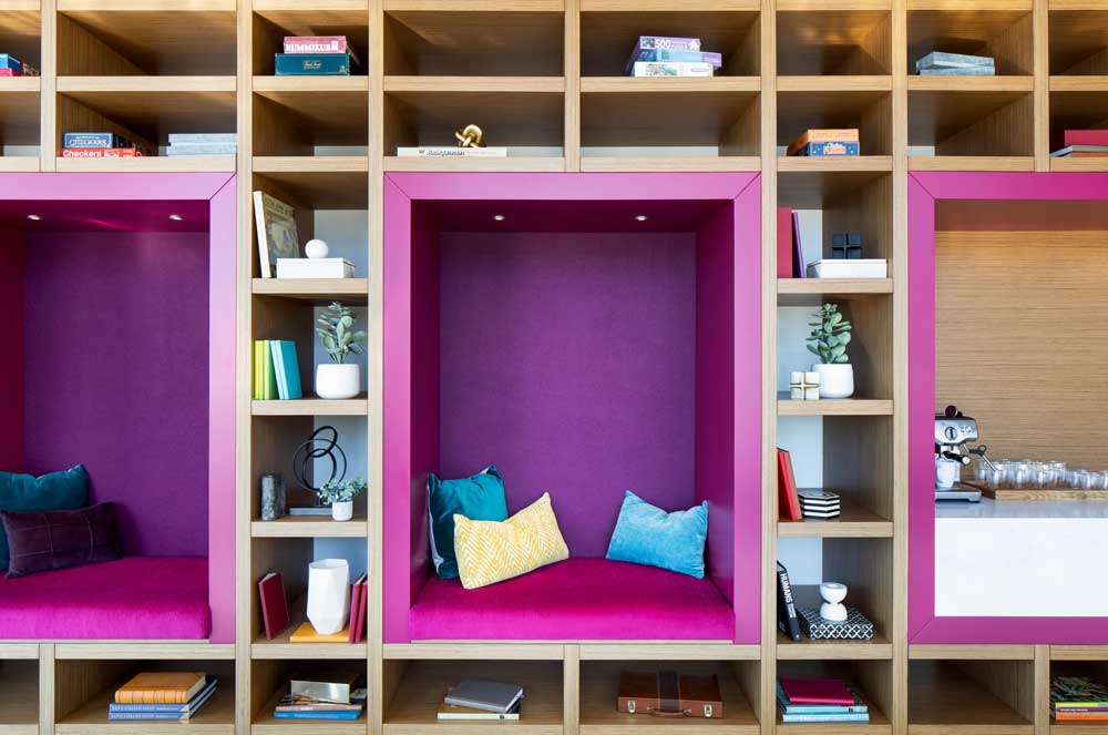 Magenta squares meant as seating booths with comfy teal coloured pillows within the built in shelving unit add a pop of colour in the space