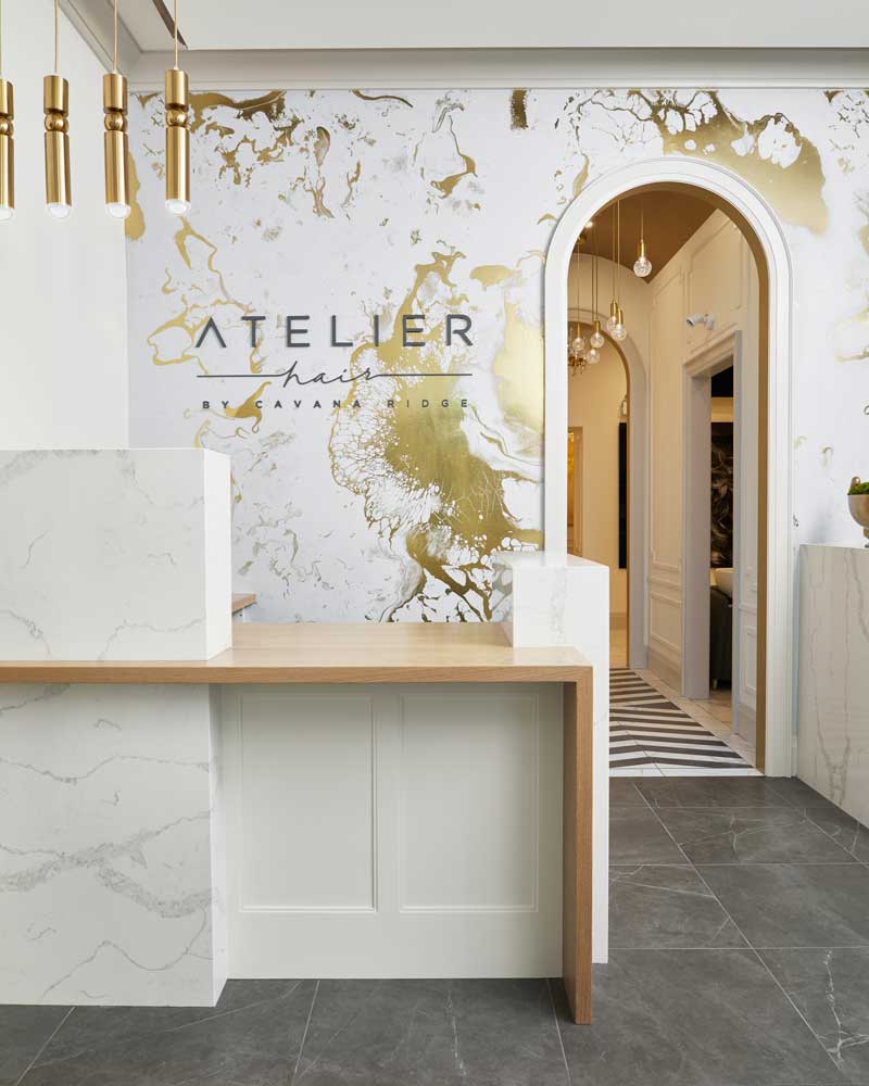 Reception desk of the Atelier Hair in simple clean white marble and wood design with the back wall in a gold and white marble effect