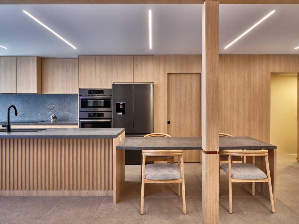 Kitchen cabinetry made of wood paneling that continues on to the surrounding walls. On the island base a wood slat material is applied and the dining table is created as an extension of the island counter