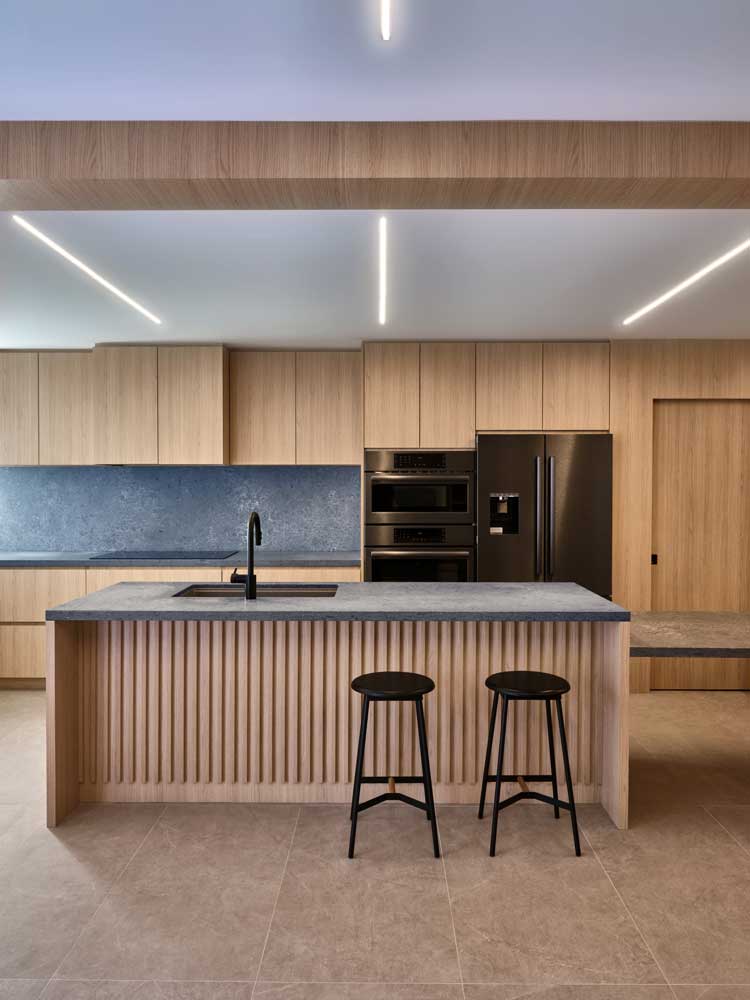 A view of the kitchen from the living room, we see the island with two black stools and in the back we see the kitchen cabinetry. Above there are thin strip LED lights running across the ceiling pointing toward the kitchen