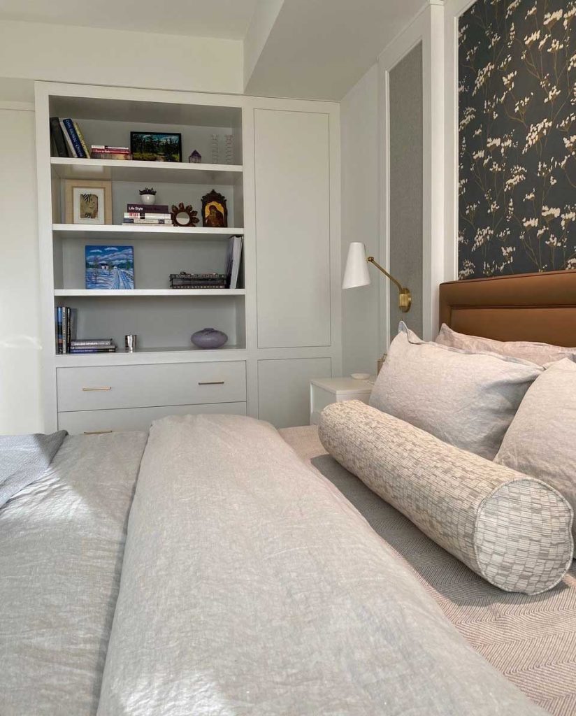Tranquil boutique hotel style bedroom, with patterned wall paper above a leatherette headboard in a brown natural colour and lighting on each side of the bed inside a paneling wall detail. A built in shelf is visible by the entrance to the bedroom