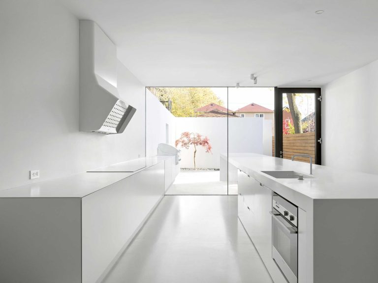 A view from the white minimalistic kitchen toward the courtyard with one Japanese Maple and the counter for dining or work that appears to project into the exterior courtyard through the floor to ceiling window.