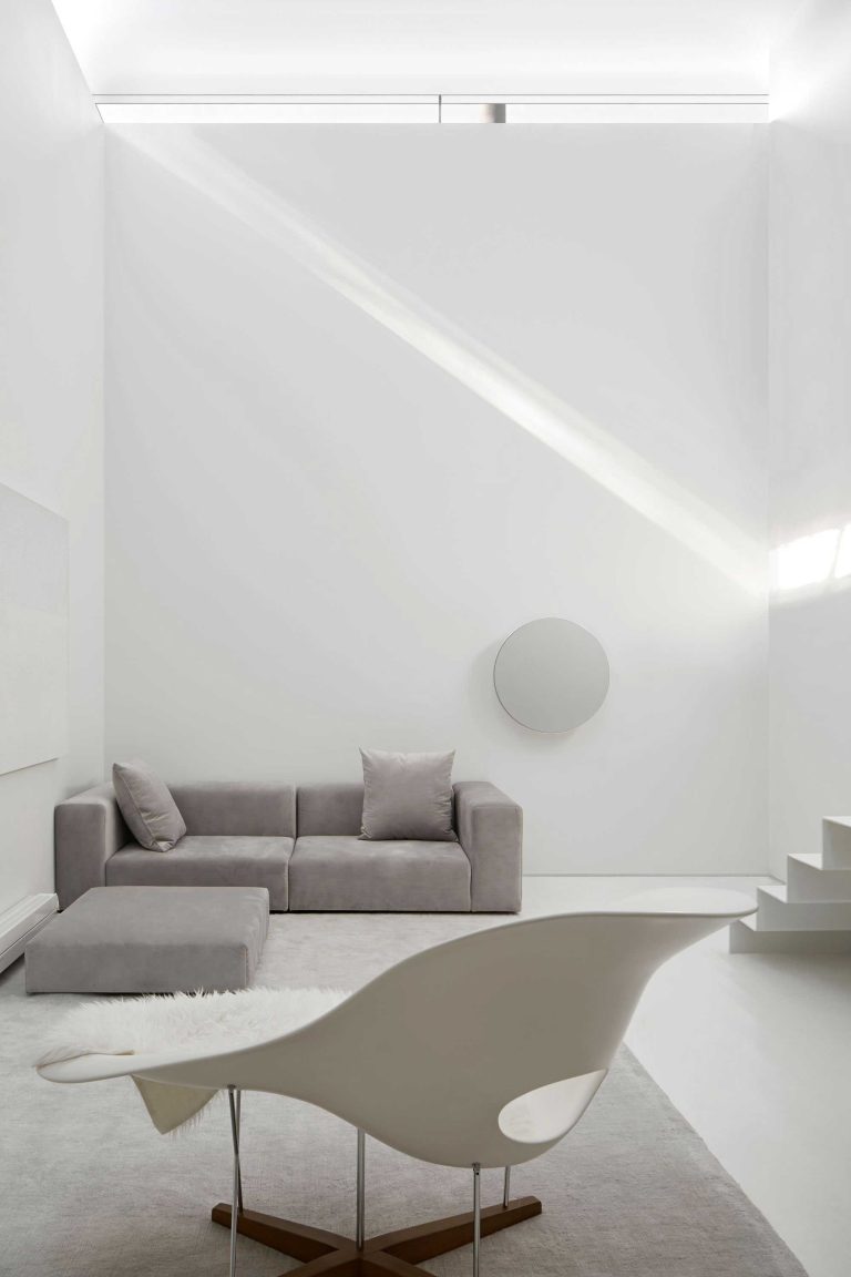 Living area on the lower level with minimalistic decor, statement chair in the foreground and a grey sofa and ottoman in the background, and a circular art piece on the tall white wall with light and shadows dancing across it