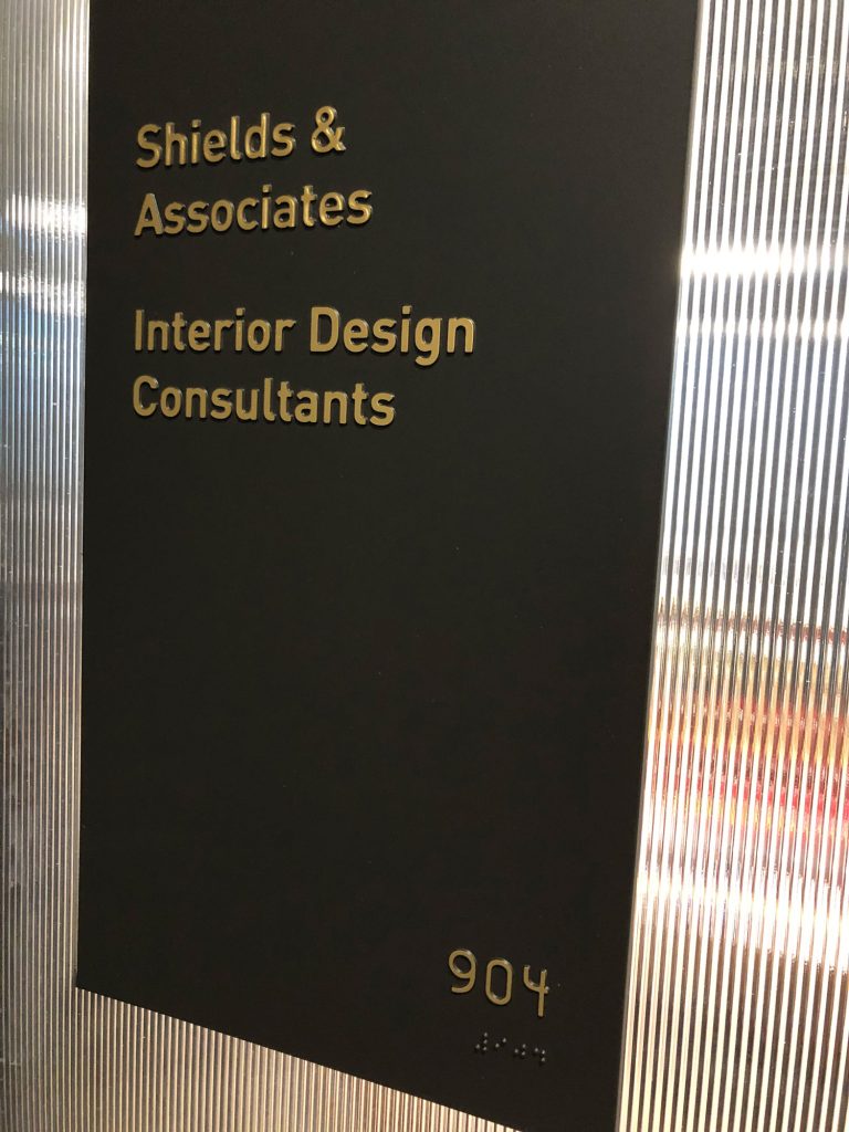 Tactile signage outside a business which reads “Shields & Associates Interior Design Consultants” with the suite number in raised lettering and braille. 
