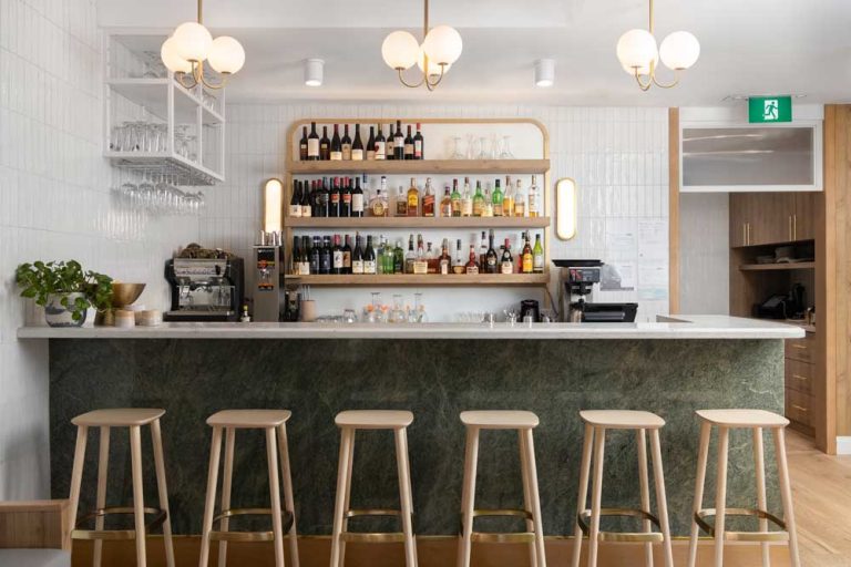 Bar with a soft green base decorated with simple bar stools. The back wall is simple and white to showcase the shelving with colourful drink bottles