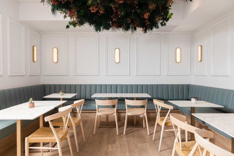 Another dining area with soft green upholstered benches along the white walls with simple white tables and light wooden chairs in front of them and greenery hanging from the ceiling