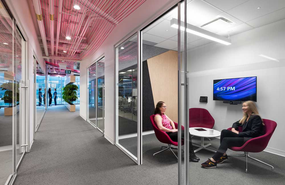 A small collaboration space behind a glass wall is visible from the hallway and corded installation above, visually leading the way into the work station area