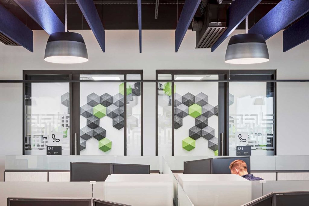 A worker at a cluster of desks in front of two private phone rooms with walls covered in hexagonal acoustical paneling.