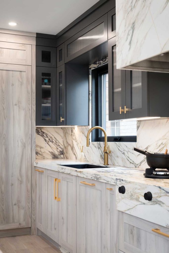 Kitchen countertops and backsplash are covered in luxurious marble  with black upper cabinets and light wood finish on the bottom cabinetry. Gold finish hardware throughout