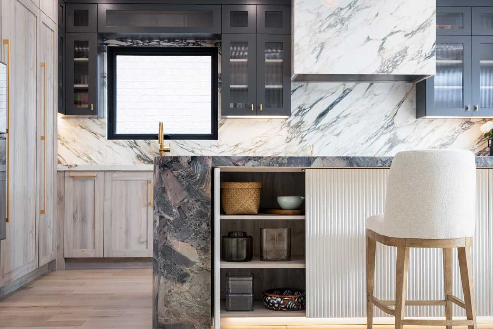 Kitchen countertops and backsplash are covered in luxurious marble  with black upper cabinets and light wood finish on the bottom cabinetry. Island covered in a darker coloured marble and a storage cabinet underneath