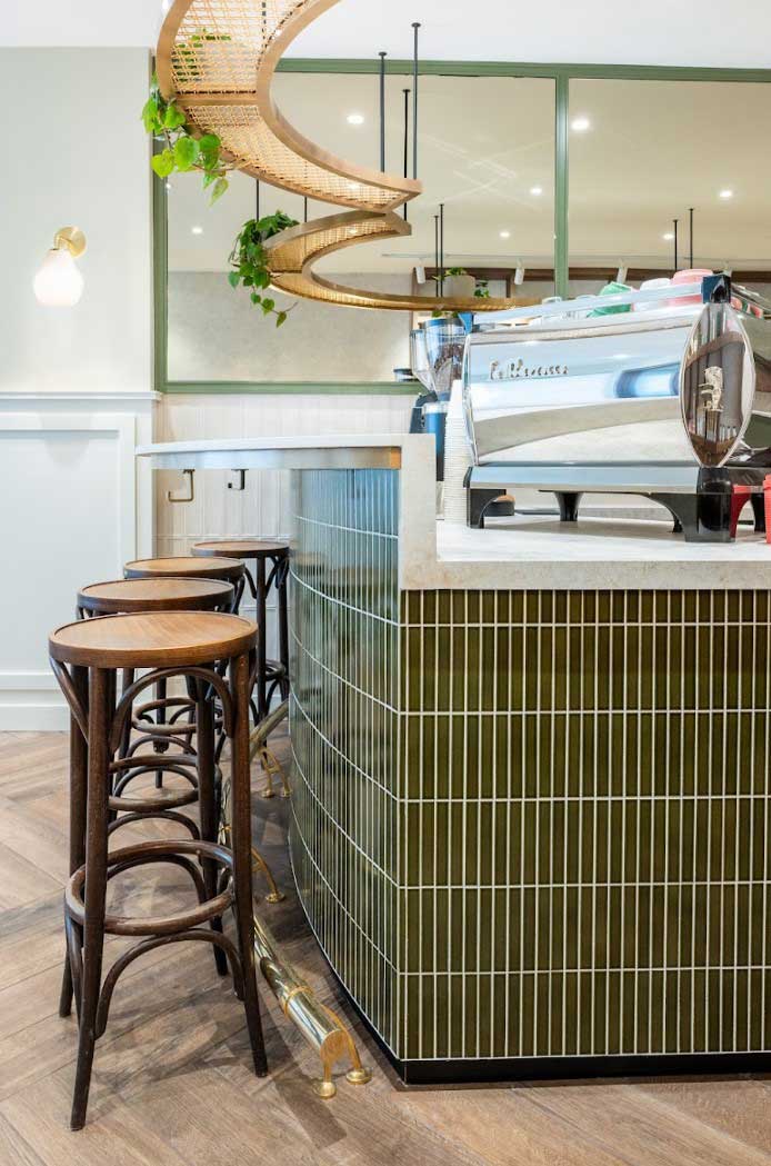 Detail of cafe counter , green tile on the counter base and wooden bar stools. Mirror in the back wall makes the space seem brighter and larger and plants haging from the overhead curved detail