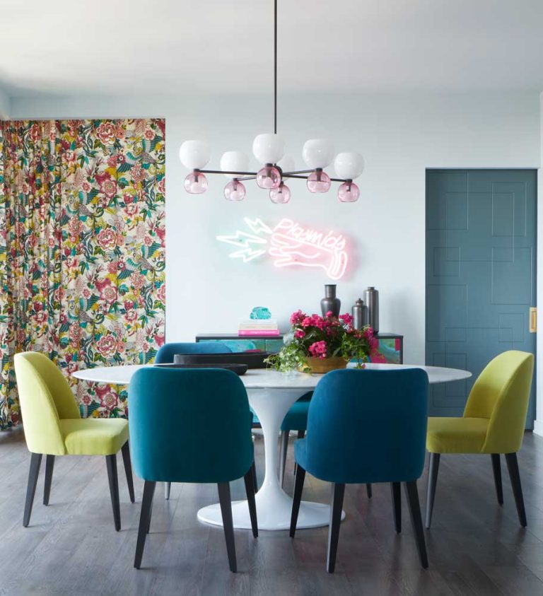 Teal and neon yellow dining chairs around a mid century modern white dining table and statement lighting above. Behind we can see the sliding door to the bedroom and neon sign on the wall above the sideboard