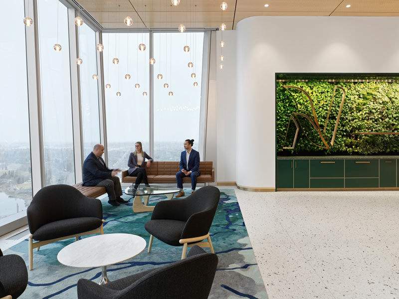 The waiting area by the reception desk is grounded by a carpet resembling water and a biophilic detail on the wall to the right, while on the left the floor to ceiling windows provide views of the city
