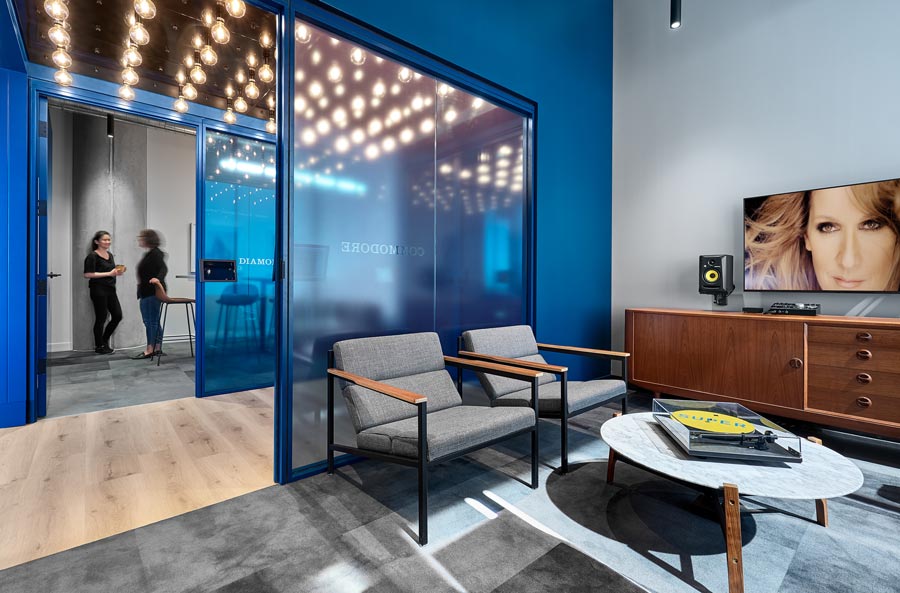 A fun lounge area with a blue accent wall and mid century inspired furniture. Outside the room in a hallway lit by large bulb lights along the ceiling