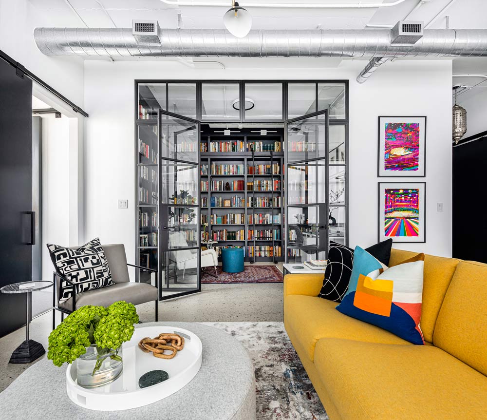 A view from the living area through large glass doors with black frames into the library with colourful book collection on custom-designed shelves