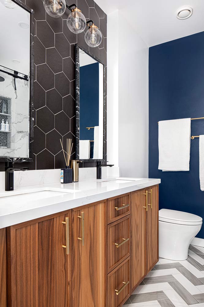 Bathroom with wood finish vanity, black hexagon tile on the wall above and industrial vanity lighting, while blue accent wall peeks on the side