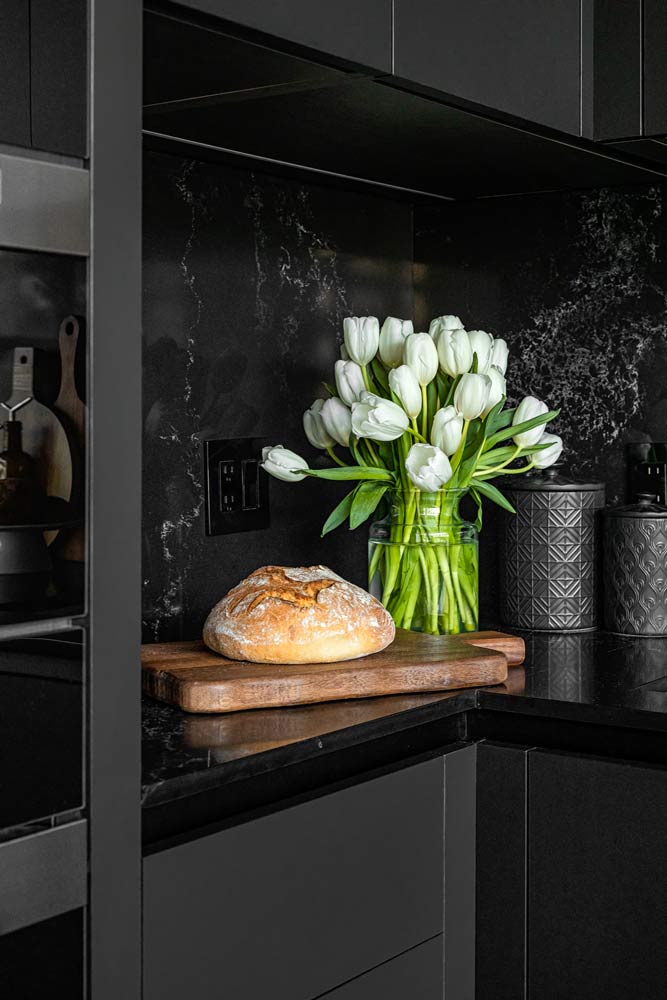 Black matte kitchen cabinets without hardware and matching black stone on counters and backsplash. Bread and flowers as display details are on the counter