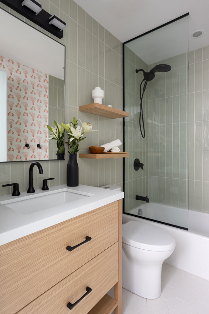 A view of the smaller bathroom toward the tub, with a wood coloured vanity against the sage green wall tile and the red and white fun patterned wall paper on the opposite wall visible in the mirror. The hardware and mirror are industrial style with black finishes