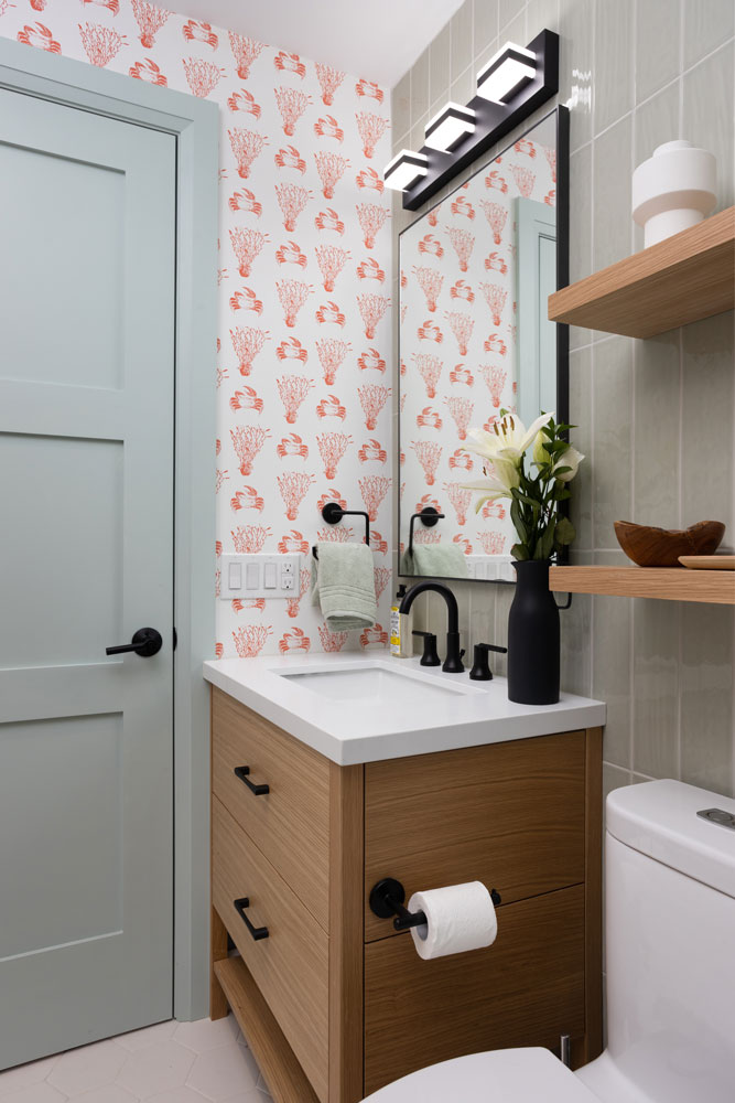 A detail of the smaller bathroom with a wood coloured vanity against a fun red and white patterned wall paper on the side wall and sage coloured vertical tile on the front wall. The hardware and mirror are industrial style with black finishes