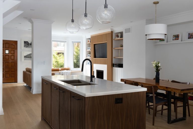 A view of the open concept kitchen, dining and living area in a predominantly white and neutral colour scheme with wooden accents throughout. Fire place in the living room at the back of the photo has a vertical wooden slat detail