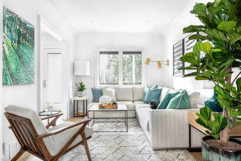 The grand view of the living room as seen from the dining room toward the big window at the other end of the living room looking onto to the street. A light and bright room in neutral tones with green and aqua accents and a large plant on the right, between the living and dining room.