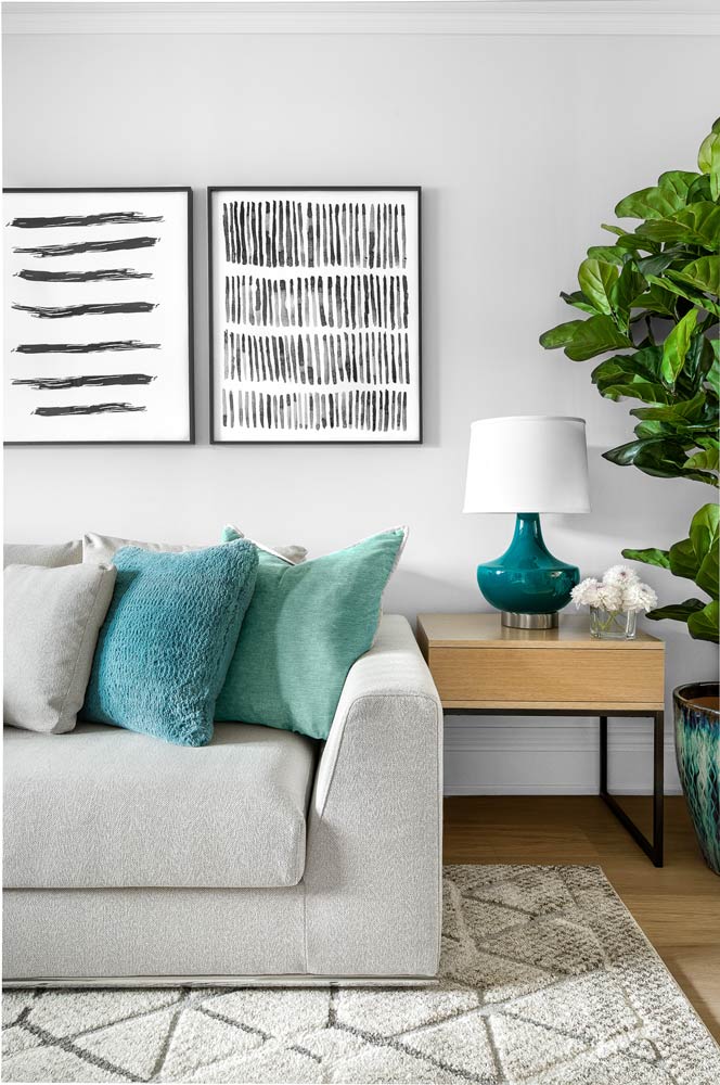 A vignette showing the living room counch and side table with a lamp in a vibrant aqua blue and modern black and white art work on the wall. The pillows on the sofa are also in tones of aqua and go with the lamp