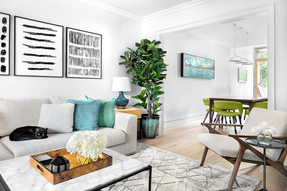 Mid century inspired living room in neutral tones with aqua accents in pillows and lamp, and in the background the dining room is visible with aqua and green accents, like dining chairs and artwork.